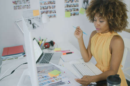 Elegant ethnic woman sitting at working desktop with gadgets and looking through papers while working in stylish office.の写真素材