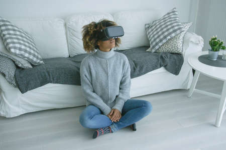 Young ethnic model in sweater sitting on floor of modern living room and wearing goggles of virtual reality experiencing another world.の写真素材