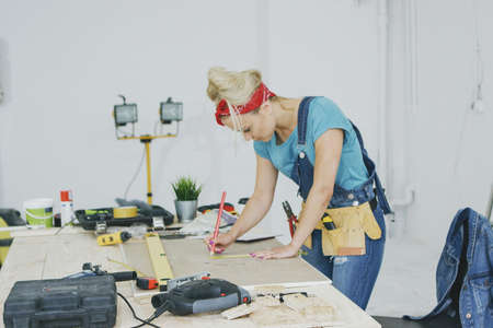 Side view of young blond woman in jeans overalls and tool belt bending over wooden workbench drawing lines with pencil on plywood surrounded by instruments in workshop.の写真素材
