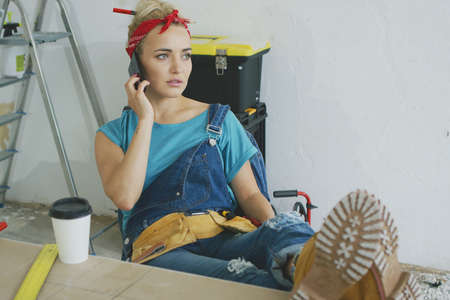Young blond woman in jeans overalls sitting with legs on top of wooden desk with tools and talking...