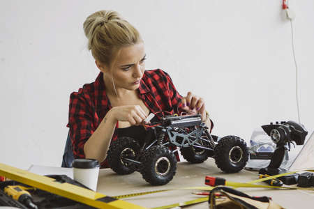 Beautiful young woman in casual checkered shirt sitting at wooden workbench with tools and fixing...
