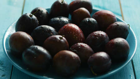 Closeup washed purple plumbs with water drops in plate on blue wooden backgroundの写真素材