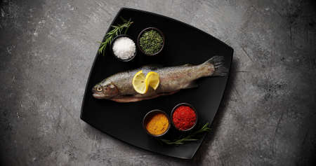 From above shot of ceramic plate with assorted aromatic spices and fresh uncooked trout standing on...
