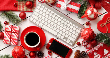 From above shot of assorted Christmas decorations and cute gift boxes lying on white lumber tabletop around modern devices and cup of fresh hot drinkの写真素材