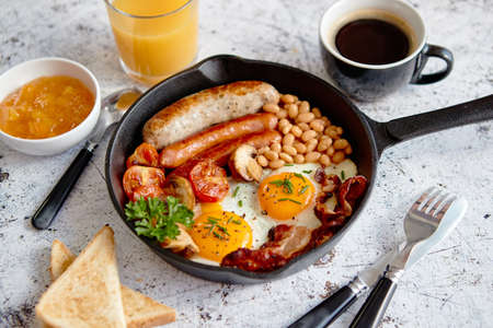 English breakfast in iron cooking pan with fried eggs, sausages, bacon, beaked beans, toasts, orange juice and black coffee on stone background.の写真素材