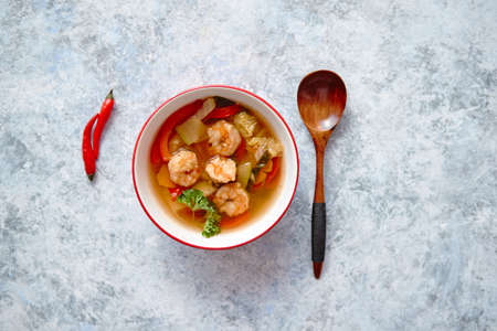 Traditional Tom Yum spicy Thai soup with shrimp, seafood, coconut milk and chili pepper in served red bowl. Fresh chilli pepper and wooden spoon on sides. Top view with copy space.の写真素材