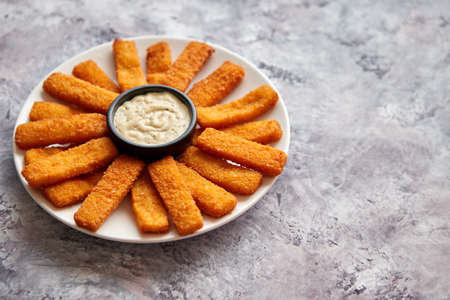 Crumbed fish sticks served with garlic dip sauce on a white plate on a stone table. Top view with...