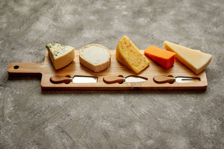 Assortment of various kinds of cheeses served on wooden board with fork and knives. Placed on...