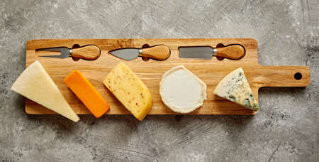 Assortment of various kinds of cheeses served on wooden board with fork and knives. Placed on...