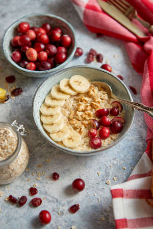 Ceramic bowl of oatmeal porridge with banana, fresh cranberries and walnuts on stone table top view in flat lay style. Healthy breakfast and diet food.の写真素材