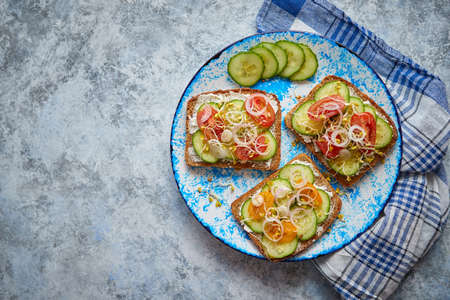 Plate with healthy toasts with cucomber, cherry tomatoes, crumbled feta cheese and radish sprouts on a black plate. Table top view. Gray stone background.の写真素材