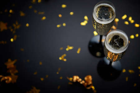 Two glasses full of sparkling champagne wine with golden decoration of confetti and serpentines on black elegant background. Top view with copy space. Festive or party concept.の写真素材
