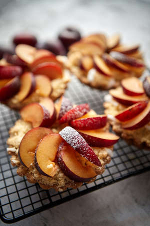 Close up of homemade crumble tarts with fresh plum slices placed on iron baking grill. Top lay on gray stone background with some whole plums, icing sugar, spoons on sideの写真素材
