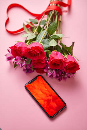 Pink roses bouquet with hart shaped bow and mobile phone with copy space screen. Placed over pink table background. Top view shot.の写真素材