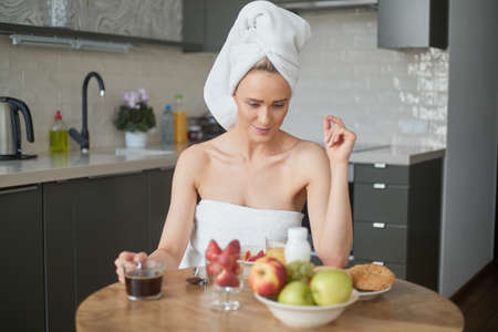 Beautiful middle aged woman sitting in her kitchen in the morning just after shower. She is wrapped in towels. Eating healthy breakfast.の写真素材