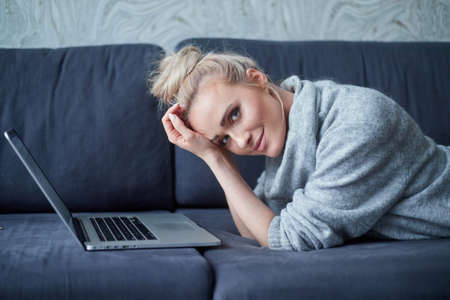 Close up Happy Pretty Young Woman Wearing Casual Clothing. Lying Prone on Sofa with Laptop While Looking at the Camera.の写真素材