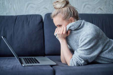 Young blond woman lying on sofa with face hidden in sweater and looking at laptop screen.の写真素材
