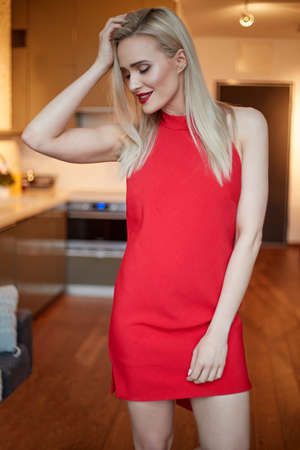 Stylish elegant blonde woman in home living room, wearing red sexy dress. Posing to the camera.の写真素材