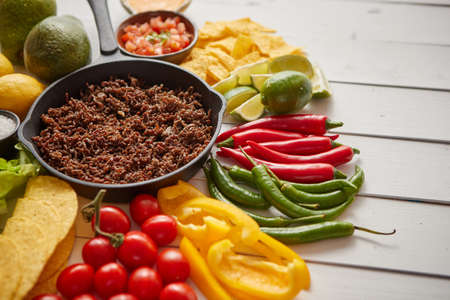 Chili con carne in frying pan on white wooden table. Ingredients for making Chili con carne.Top view. Chili with meat, nachos, tacos, limes, avocado, hot pepper. Mexican Texas traditional dishの写真素材
