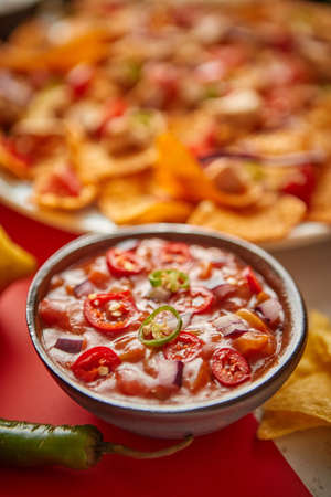 Close up on hot tomato dip in ceramic bowl with various freshly made Mexican foods assortment in the back. Placed on colorful table. With nachos, tacos, tortillas, grilled meat, dips, salsa and vegetables.の写真素材