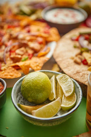 Close up on lime slices in ceramic bowl with various freshly made Mexican foods assortment in the back. Placed on colorful table. With nachos, tacos, tortillas, grilled meat, dips, salsa and vegetables.の写真素材