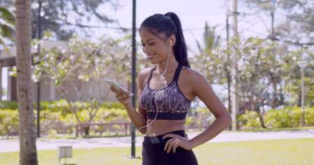 Sportive slim Asian woman in earphones surfing mobile phone in hand while having break during workout in park Thailand, Phuketの写真素材