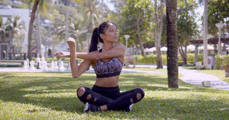 Slim ethnic woman in fitness clothing sitting in lotus position with crossed legs and warming up arms in green tropical park in Thailand, Phuketの写真素材