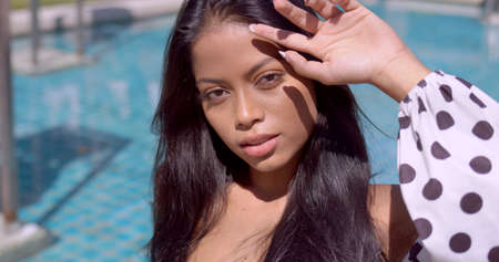 High angle of crop gorgeous Asian female in white and brown polka dot clothes, with raised hand looking with sensual gaze at camera against swimming pool in sunny day in Thailandの写真素材
