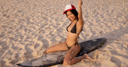 Happy smiling brunette with slim tanned body wearing bikini and cap sitting on surfboard on sandy beach and having fun pouring sand from handの写真素材