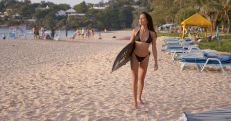 Active slim brunette in black bikini walking on sandy beach with surfboard under arm and looking at sea while preparing for surfingの写真素材