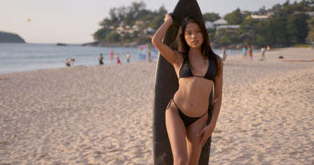 Relaxed young female in black bikini enjoying summer day on tropical seaside in Phuket and standing on sandy beach with surfboardの写真素材
