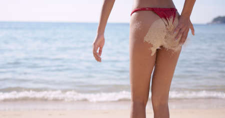 Crop back view woman in red bikini with perfect tanned body in water drops on skin and sand on buttocks, standing on tropical seaside in Phuket Thailandの写真素材