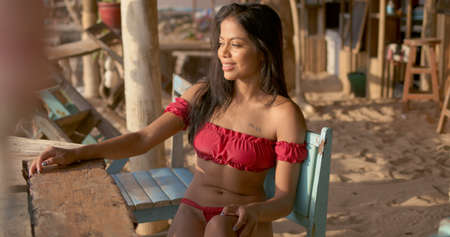Side view of relaxed smiling brunette with tanned skin and in trendy red bikini sitting at wooden table in beach cafe and enjoying summer holidays, in Phuket Thailandの写真素材