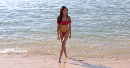 Attractive ethnic young woman with long dark hair wearing stylish red bikini standing on tropical sandy beach with clear sea water in Phuket Thailandの写真素材