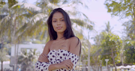 Long haired hot Asian woman in dress with open shoulders crossed hands and looking at camera in park in Thailand, Phuketの写真素材