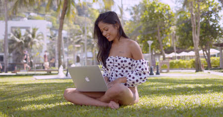 Long haired cheerful Asian woman in top with open shoulders working on laptop sitting with crossed legs in park in Thailand, Phuketの写真素材