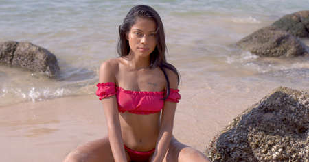 Sensual Asian lady in stylish pink bikini relaxing while sitting on seashore and looking at camera at Phuketの写真素材