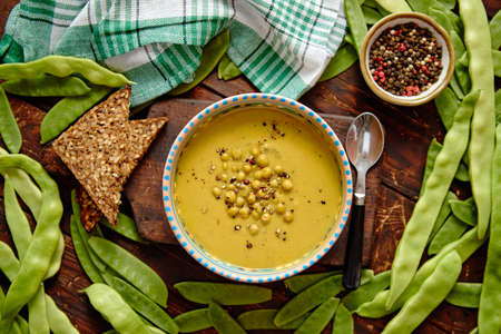Green vegetables cream soup in grey bowl placed among fresh peas and bean pods with full grain bread on wooden background. Top view with copy space.の写真素材