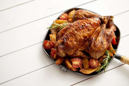 Roasted whole chicken in cast iron black pan. With potatoes, tomatos and rosemary herb. Placed in white wooden table. View from above. Shot with copy space.の写真素材