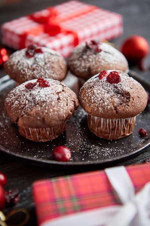 Christmas chocolate delicious muffins served on black ceramic plate. Sprinkled with powder sugar. Cranberries on top. Xmas gifts and decorations on sides.の写真素材