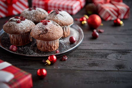 Christmas chocolate delicious muffins served on black ceramic plate. Sprinkled with powder sugar. Cranberries on top. Xmas gifts and decorations on sides.の写真素材