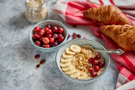 Ceramic bowl of oatmeal porridge with banana, fresh cranberries and walnuts on stone table top view in flat lay style. Healthy breakfast and diet food.の写真素材