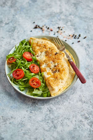 Classic egg omelette served with cherry tomato and arugula salad on side. Placed on white ceramic plate. Stone background with copy space.の写真素材