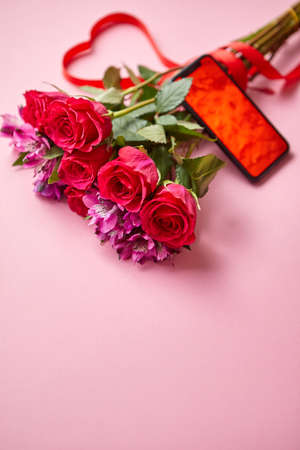 Pink roses bouquet with hart shaped bow and mobile phone with copy space screen. Placed over pink table background. Top view shot.の写真素材