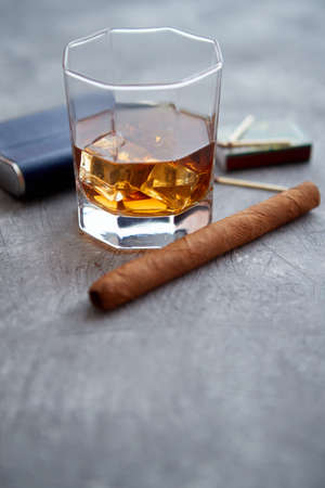 Glass of whiskey with ice, big cuban cigar, matches and hip flask placed on gray stone table.の写真素材