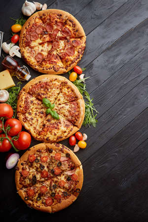 Freshly served, three various pizzas placed among tasty ingredients. Top view. Black wooden background with copy space for text.の写真素材