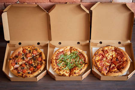 Three different kind of pizzas in delivery boxes. Placed in a row on brown wooden background. View...