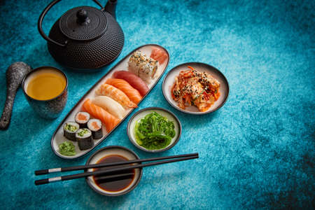 Various sushi rolls placed on ceramic plates. Traditional iron tea pot and green tea in cup. Kimchi and goma wakame salads. Soy souce and chopsticks on sides. Blue background with copy space.の写真素材