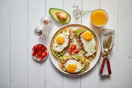 Delicious healthy breakfast with sliced avocado sandwiches with fried egg on top of bread. With orange juice, cherry tomatoes, radish sprouts, salt and peper. Flat lay, top view.の写真素材