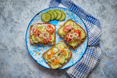 Plate with healthy toasts with cucomber, cherry tomatoes, crumbled feta cheese and radish sprouts on a black plate. Table top view. Gray stone background.の写真素材